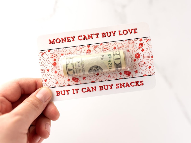 Puede incluir: Una tarjeta blanca con texto rojo que dice "El dinero no puede comprar el amor, pero puede comprar bocadillos." Se puede ver un billete de d&oacute;lar enrollado en la tarjeta.