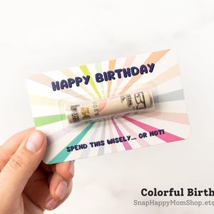 Może przedstawiać: Prostokątna kartka urodzinowa z kolorowym wzorem promieni słonecznych w pastelowych odcieniach. Tekst brzmi HAPPY BIRTHDAY na g&oacute;rze i SPEND THIS WISELY... OR NOT! na dole. Zrolowany banknot pięćdziesięciodolarowy znajduje się w plastikowej kapsule.