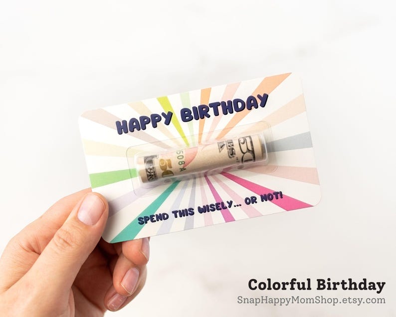 Może przedstawiać: Prostokątna kartka urodzinowa z kolorowym wzorem promieni słonecznych w pastelowych odcieniach. Tekst brzmi HAPPY BIRTHDAY na g&oacute;rze i SPEND THIS WISELY... OR NOT! na dole. Zrolowany banknot pięćdziesięciodolarowy znajduje się w plastikowej kapsule.