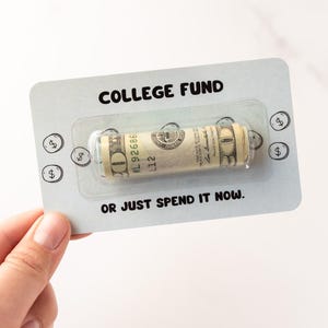 Może przedstawiać: Prostokątna jasnoszara karta z napisem COLLEGE FUND u g&oacute;ry i OR JUST SPEND IT NOW u dołu. Przezroczyste plastikowe opakowanie typu blister pośrodku zawiera zwinięty banknot dwudziestodolarowy. Małe ilustracje monet otaczają środkową część karty.