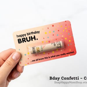 Może przedstawiać: Prostokątna karta w kolorze koralowym z wzorami konfetti, zawierająca tekst happy birthday BRUH. oraz ...we all know this is what you really wanted. Zrolowany banknot pięćdziesięciodolarowy jest zamknięty w przezroczystej plastikowej kapsule.