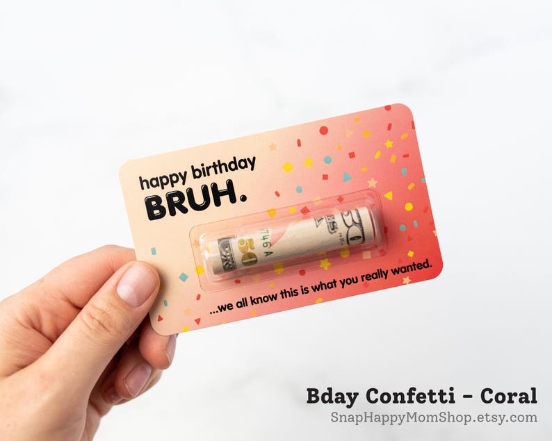 Może przedstawiać: Prostokątna karta w kolorze koralowym z wzorami konfetti, zawierająca tekst happy birthday BRUH. oraz ...we all know this is what you really wanted. Zrolowany banknot pięćdziesięciodolarowy jest zamknięty w przezroczystej plastikowej kapsule.