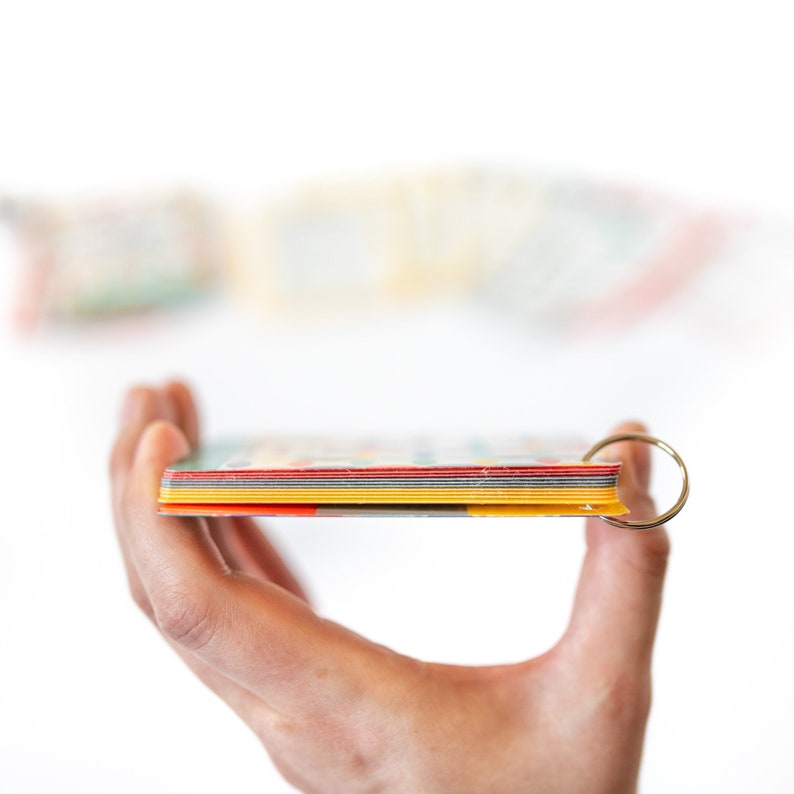 Op de afbeelding: Een stapel kleurrijke indexkaarten met afgeronde hoeken, vastgehouden in een hand. De kaarten zijn aan elkaar gebonden met een zilveren ring.