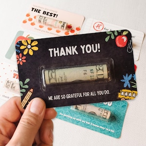 May include: A black card with the text "THANK YOU!" and a rolled up dollar bill inside a clear plastic sleeve. The card also has a colorful design with flowers, pencils, and a school bus. The text "WE ARE SO GRATEFUL FOR ALL YOU DO." is printed below the dollar bill.