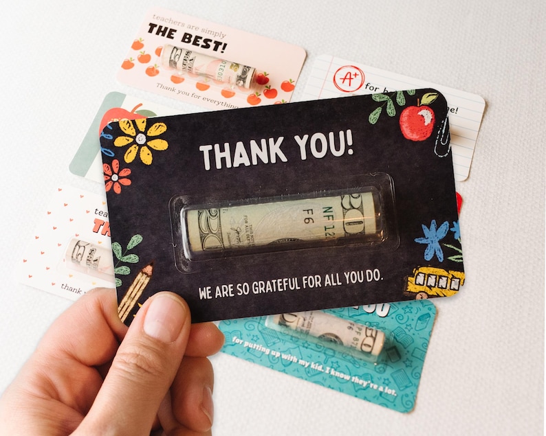 May include: A black card with the text "THANK YOU!" and a rolled up dollar bill inside a clear plastic sleeve. The card also has a colorful design with flowers, pencils, and a school bus. The text "WE ARE SO GRATEFUL FOR ALL YOU DO." is printed below the dollar bill.