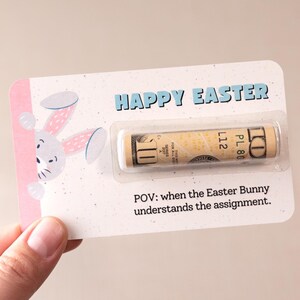 May include: A pink and white card with a cartoon bunny peeking out from the side. The card says "Happy Easter" and has a $1 bill inside. The text below the bill says "POV: when the Easter Bunny understands the assignment."