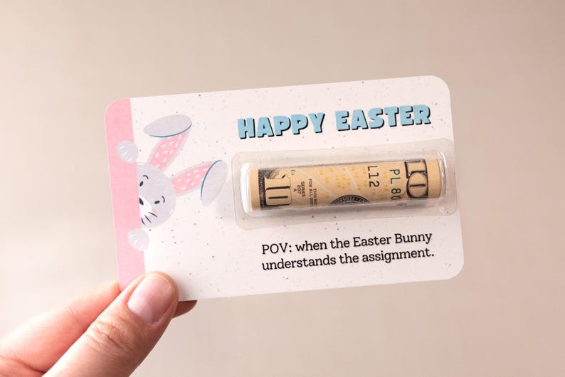 May include: A pink and white card with a cartoon bunny peeking out from the side. The card says "Happy Easter" and has a $1 bill inside. The text below the bill says "POV: when the Easter Bunny understands the assignment."