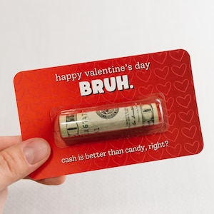 Puede incluir: Tarjeta roja con corazones blancos y el texto "happy valentine's day BRUH. cash is better than candy, right?" Un billete de d&oacute;lar enrollado es visible en el centro de la tarjeta.