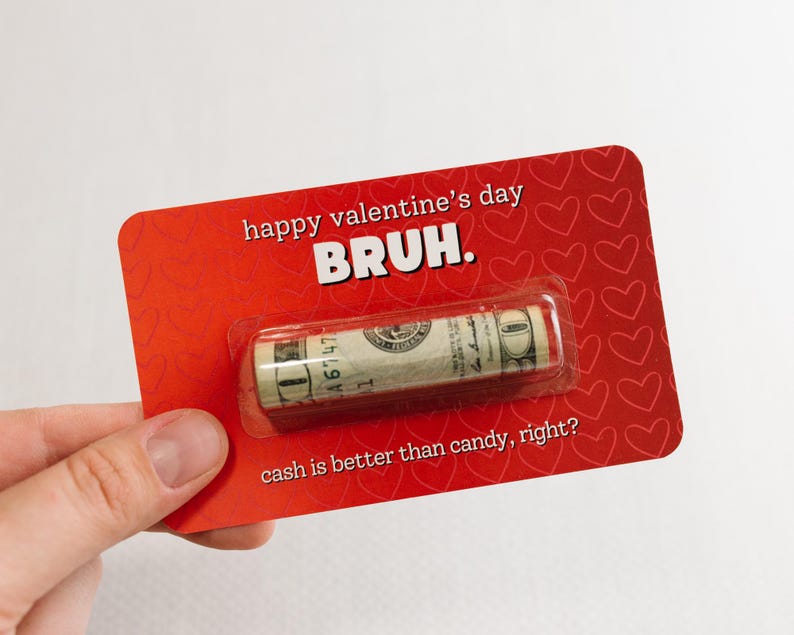 Puede incluir: Tarjeta roja con corazones blancos y el texto "happy valentine's day BRUH. cash is better than candy, right?" Un billete de d&oacute;lar enrollado es visible en el centro de la tarjeta.