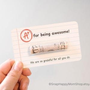 May include: A white card with red and black text that reads "A+ for being awesome!" with a rolled up dollar bill inside. The text "We are so grateful for all you do." is printed below the bill. The card has a lined background.