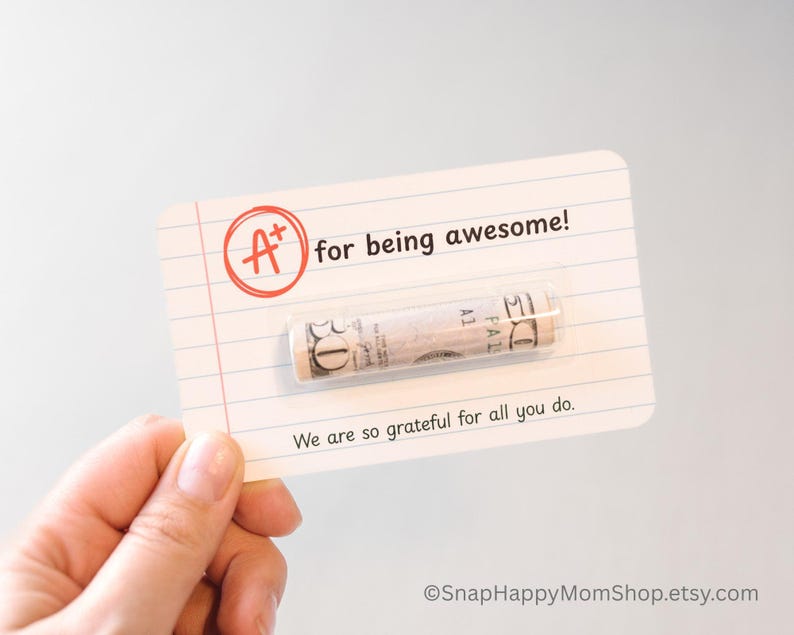 May include: A white card with red and black text that reads "A+ for being awesome!" with a rolled up dollar bill inside. The text "We are so grateful for all you do." is printed below the bill. The card has a lined background.