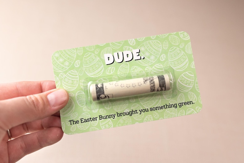 May include: A green card with a white Easter egg pattern and the text "DUDE." A rolled up dollar bill is inside a clear plastic sleeve. The text "The Easter Bunny brought you something green." is printed below the dollar bill.