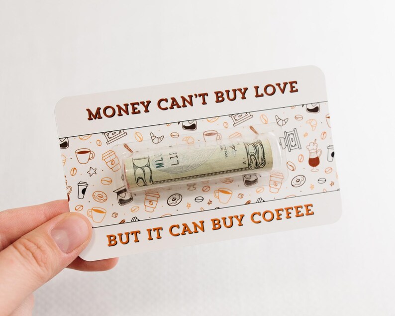 Puede incluir: Una tarjeta blanca con texto marr&oacute;n que dice "El dinero no puede comprar el amor pero puede comprar caf&eacute;." Un billete de d&oacute;lar enrollado es visible a trav&eacute;s de una ventana de pl&aacute;stico transparente en la tarjeta. La tarjeta est&aacute; decorada con un patr&oacute;n de tazas de caf&eacute;, donuts y estrellas.