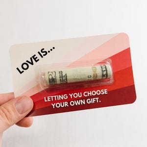 Puede incluir: Una tarjeta roja y rosa con el texto "LOVE IS... LETTING YOU CHOOSE YOUR OWN GIFT." y un billete de d&oacute;lar enrollado dentro de una manga de pl&aacute;stico transparente.