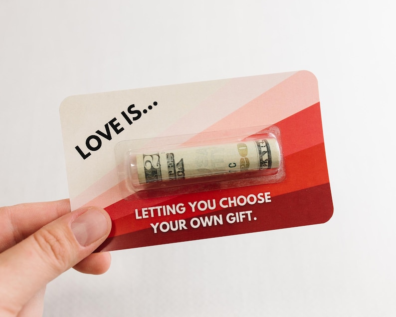 Puede incluir: Una tarjeta roja y rosa con el texto "LOVE IS... LETTING YOU CHOOSE YOUR OWN GIFT." y un billete de d&oacute;lar enrollado dentro de una manga de pl&aacute;stico transparente.