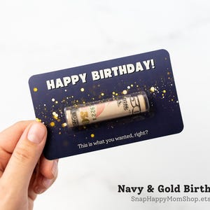Może przedstawiać: Prostokątna granatowa kartka urodzinowa ze złotymi akcentami brokatu oraz napisem HAPPY BIRTHDAY! i This is what you wanted, right? Przezroczyste plastikowe opakowanie typu blister zawiera zwinięty banknot pięćdziesięciodolarowy.