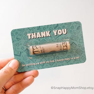 May include: A teal card with a white border and a rolled up dollar bill inside a clear plastic window. The card has a pattern of books and stars and says "THANK YOU" in white letters. The text below the window says "for putting up with my kid. I know they're a lot."