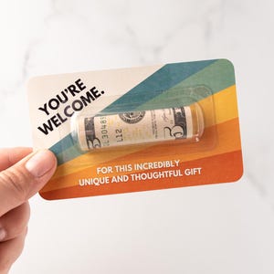 Może przedstawiać: Prostokątna karta z wzorem tęczowych pask&oacute;w w stylu retro, zawierająca tekst YOU'RE WELCOME oraz FOR THIS INCREDIBLY UNIQUE AND THOUGHTFUL GIFT. Przezroczyste plastikowe opakowanie typu blister zawiera zwinięty banknot pięciodolarowy.