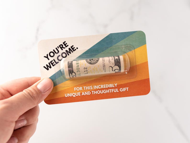Może przedstawiać: Prostokątna karta z wzorem tęczowych pask&oacute;w w stylu retro, zawierająca tekst YOU'RE WELCOME oraz FOR THIS INCREDIBLY UNIQUE AND THOUGHTFUL GIFT. Przezroczyste plastikowe opakowanie typu blister zawiera zwinięty banknot pięciodolarowy.
