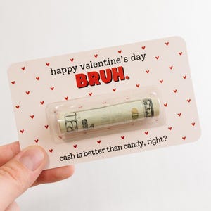 Puede incluir: Tarjeta rosa con corazones rojos y el texto "happy valentine's day BRUH." Un billete de d&oacute;lar enrollado est&aacute; dentro de una manga de pl&aacute;stico transparente en la tarjeta. El texto "cash is better than candy, right?" est&aacute; en la parte inferior de la tarjeta.