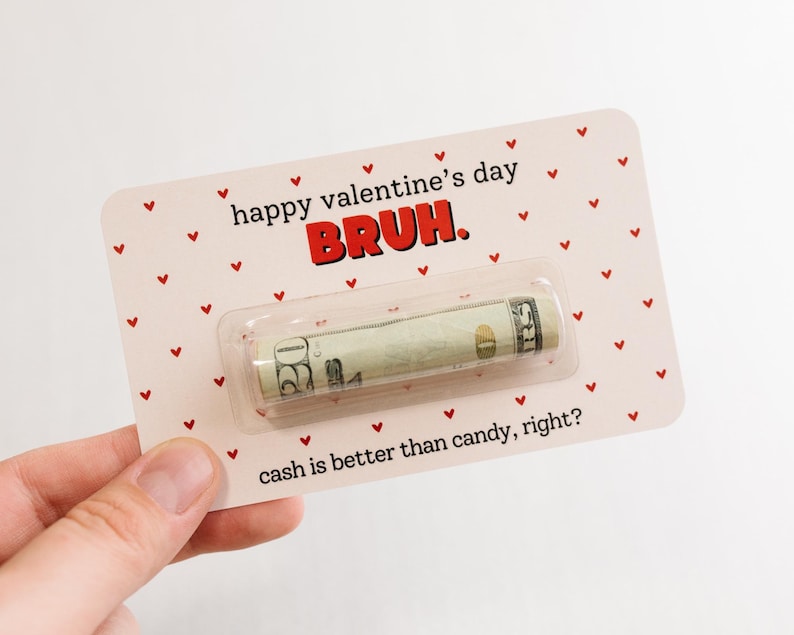 Puede incluir: Tarjeta rosa con corazones rojos y el texto "happy valentine's day BRUH." Un billete de d&oacute;lar enrollado est&aacute; dentro de una manga de pl&aacute;stico transparente en la tarjeta. El texto "cash is better than candy, right?" est&aacute; en la parte inferior de la tarjeta.