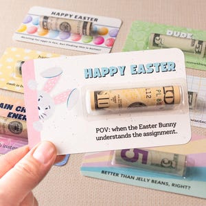 May include: A set of colorful Easter cards with a bunny rabbit illustration and the text "Happy Easter". Each card features a rolled-up dollar bill inside a clear plastic sleeve. The text "POV: when the Easter Bunny understands the assignment." is printed on one of the cards.
