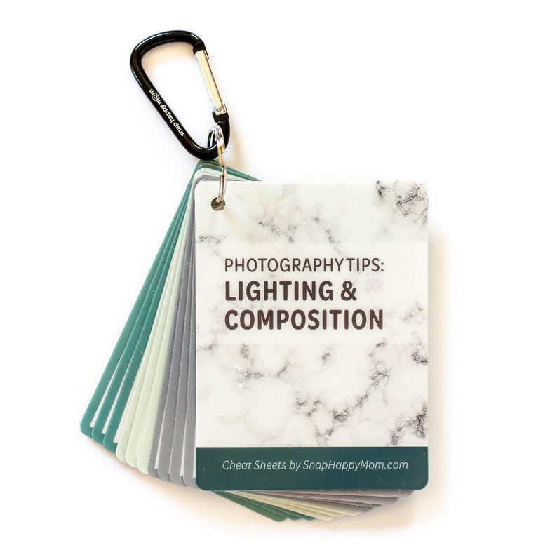 May include: A set of photography cheat sheets with a marble pattern and the text "PHOTOGRAPHY TIPS: LIGHTING & COMPOSITION" and "Cheat Sheets by SnapHappyMom.com". The cheat sheets are attached to a black carabiner.