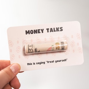 Puede incluir: Una tarjeta rosa con el texto "MONEY TALKS" y un billete de $ 20 enrollado dentro de una ventana de pl&aacute;stico transparente. El texto "this is saying "treat yourself" est&aacute; impreso debajo del billete.