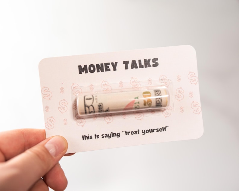 Puede incluir: Una tarjeta rosa con el texto "MONEY TALKS" y un billete de $ 20 enrollado dentro de una ventana de pl&aacute;stico transparente. El texto "this is saying "treat yourself" est&aacute; impreso debajo del billete.