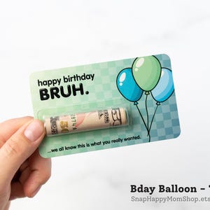 Może przedstawiać: Prostokątna, turkusowa kartka urodzinowa w kratkę z trzema ilustrowanymi balonami oraz napisem "happy birthday BRUH." i "...we all know this is what you really wanted." Zrolowany banknot pięćdziesięciodolarowy jest zamknięty w przezroczystej plastikowej kapsule.