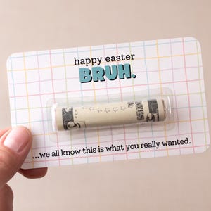 May include: A white card with a grid pattern and the text "happy easter BRUH."  A rolled up dollar bill is inside a clear plastic sleeve on the card. The text "...we all know this is what you really wanted." is at the bottom of the card.