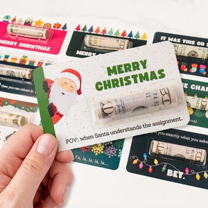 May include: A hand holding a green card with a Santa Claus illustration and the text "MERRY CHRISTMAS". Several other Christmas-themed cards with rolled-up bills are visible, featuring various designs and text.