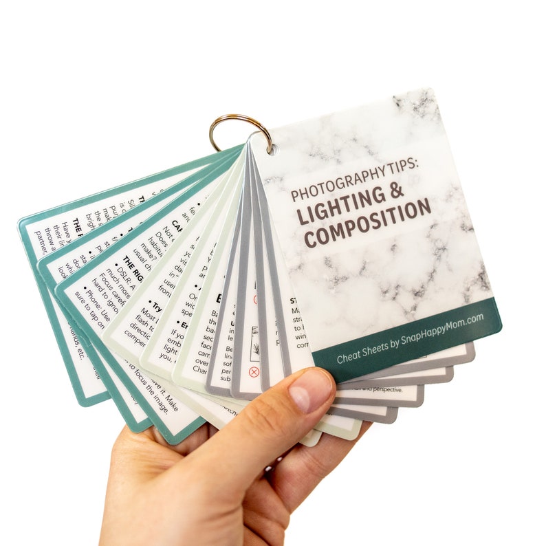 May include: A set of photography tips cheat sheets with a marble pattern background. The cheat sheets are fanned out and have titles like "The Big" and "CAF". The text "Photography Tips: Lighting & Composition" is on the top card. The text "Cheat Sheets by SnapHappyMom.com" is on the bottom card.