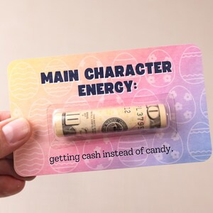 May include: A pink, blue, and yellow card with a rolled up dollar bill inside a plastic sleeve. The card says "Main Character Energy: getting cash instead of candy."