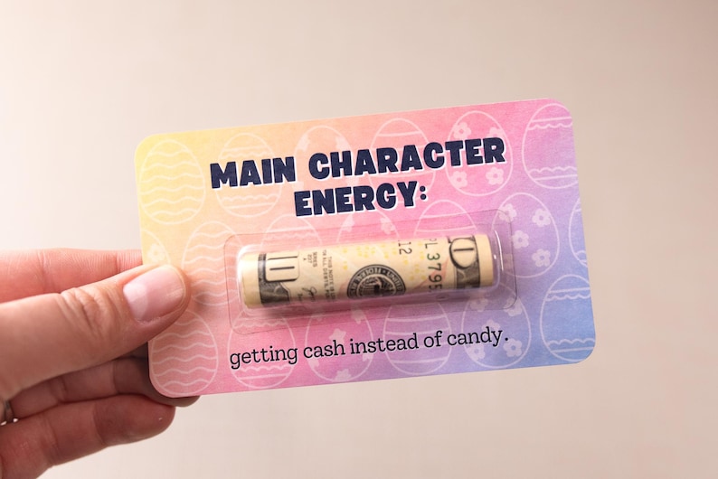 May include: A pink, blue, and yellow card with a rolled up dollar bill inside a plastic sleeve. The card says "Main Character Energy: getting cash instead of candy."