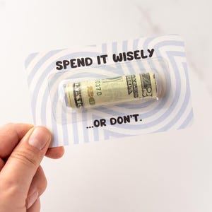 Może przedstawiać: Prostokątna karta z jasnoniebieskim i białym wzorem w okręgi, zawierająca przezroczyste plastikowe opakowanie typu blister ze zwiniętym banknotem dwudziestodolarowym. Napis SPEND IT WISELY znajduje się nad banknotem, a ...OR DON'T. pod nim.