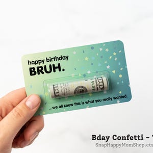 Może przedstawiać: Prostokątna kartka urodzinowa w kolorze morskim z wzorem w konfetti oraz napisem "happy birthday BRUH." i "...we all know this is what you really wanted." Przezroczyste plastikowe opakowanie typu blister zawiera zwinięty banknot studolarowy.