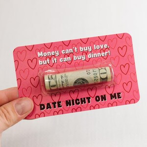 Puede incluir: Una tarjeta rosa con corazones impresos en ella. La tarjeta dice "El dinero no puede comprar el amor, pero puede comprar la cena!" y "Cena rom&aacute;ntica por mi cuenta". Un billete de d&oacute;lar enrollado est&aacute; unido a la tarjeta.