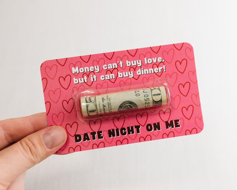 Puede incluir: Una tarjeta rosa con corazones impresos en ella. La tarjeta dice "El dinero no puede comprar el amor, pero puede comprar la cena!" y "Cena rom&aacute;ntica por mi cuenta". Un billete de d&oacute;lar enrollado est&aacute; unido a la tarjeta.