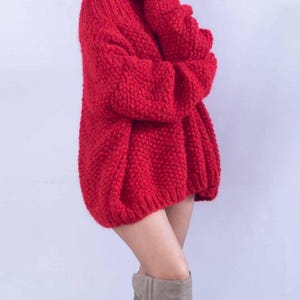 May include: A bright red, chunky knit sweater featuring a high turtleneck collar and long, loose-fitting sleeves. The garment is crafted with a thick, textured yarn in a prominent stitch pattern, providing a voluminous and relaxed silhouette.