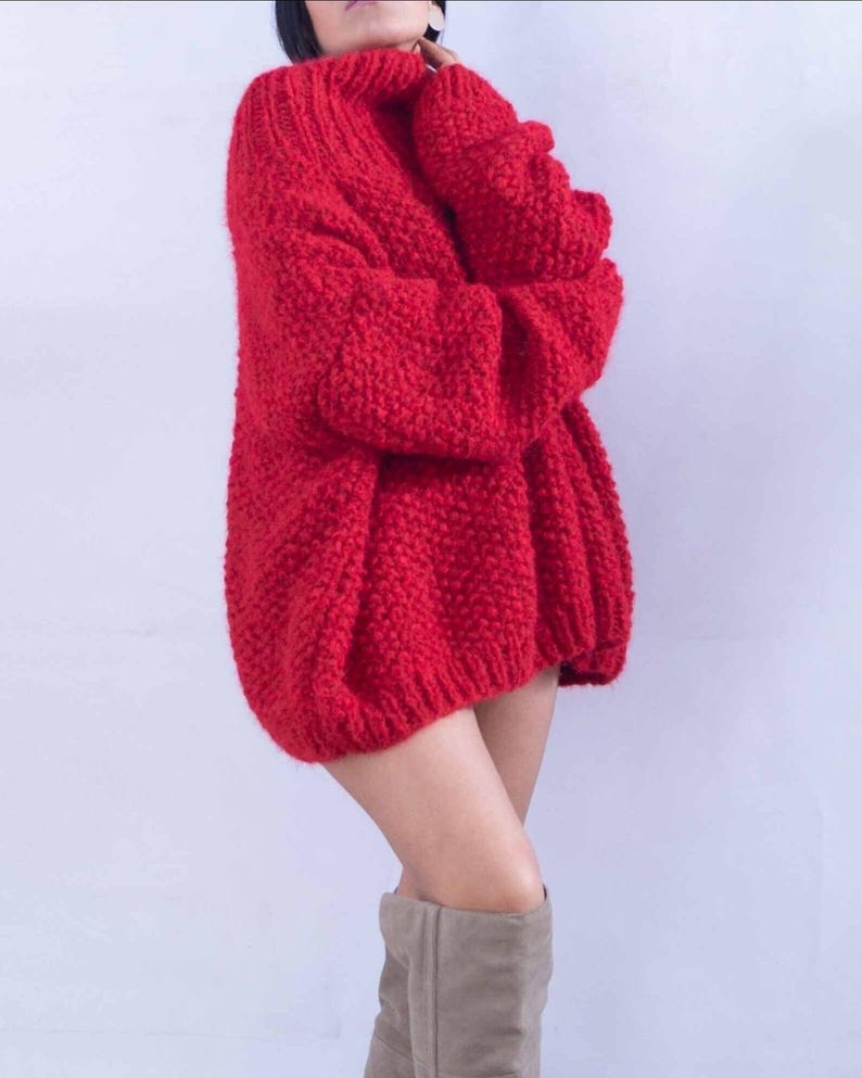 May include: A bright red, chunky knit sweater featuring a high turtleneck collar and long, loose-fitting sleeves. The garment is crafted with a thick, textured yarn in a prominent stitch pattern, providing a voluminous and relaxed silhouette.