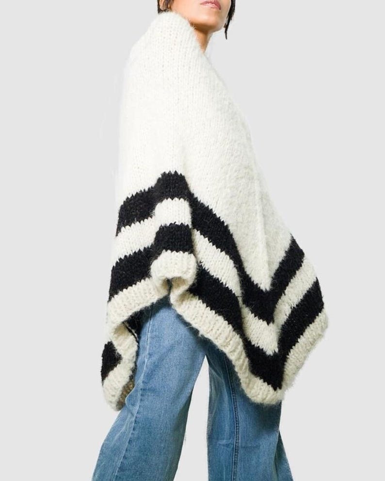 Pu&ograve; includere: Un poncho in maglia grossa color bianco sporco con due spesse strisce orizzontali nere lungo l'orlo inferiore. Il capo ha un aspetto testurizzato e soffice ed &egrave; indossato sopra un paio di jeans in denim azzurro a gamba dritta.