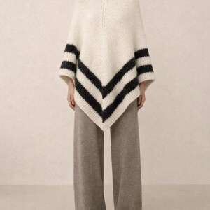 Pu&ograve; includere: Un poncho in maglia color crema con orlo a V e due strisce orizzontali nere vicino al bordo inferiore. Il capo &egrave; abbinato a pantaloni a gamba larga color tortora m&eacute;lange che arrivano fino a terra, creando un insieme dalla texture definita.