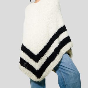 Pu&ograve; includere: Un poncho in maglia grossa color crema con due larghe strisce orizzontali nere vicino all'orlo. Il capo presenta uno scollo rotondo e una trama spessa e testurizzata, abbinato a un paio di jeans in denim azzurro visibili sotto.