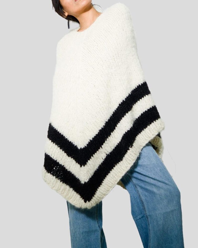 Pu&ograve; includere: Un poncho in maglia grossa color crema con due larghe strisce orizzontali nere vicino all'orlo. Il capo presenta uno scollo rotondo e una trama spessa e testurizzata, abbinato a un paio di jeans in denim azzurro visibili sotto.