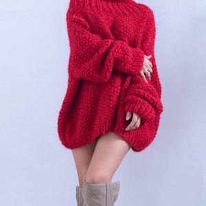 May include: A bright red oversized sweater featuring a chunky knit texture, a high mock neckline, and long sleeves with ribbed cuffs. The garment has a loose, relaxed fit that extends to the upper thigh, paired with light beige knee-high boots.