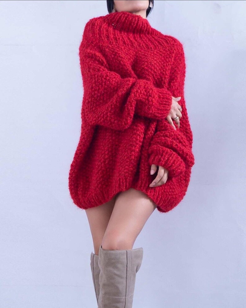 May include: A bright red oversized sweater featuring a chunky knit texture, a high mock neckline, and long sleeves with ribbed cuffs. The garment has a loose, relaxed fit that extends to the upper thigh, paired with light beige knee-high boots.