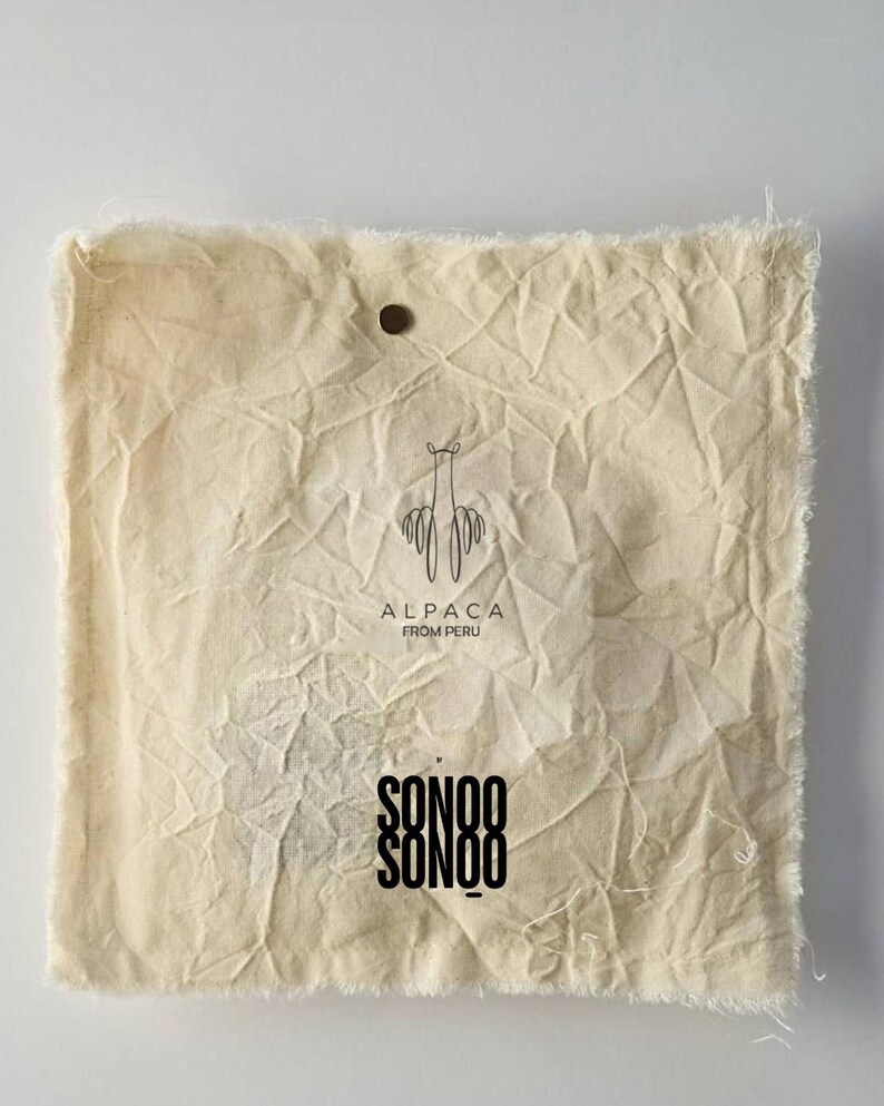 Pu&ograve; includere: Una pochette quadrata in tessuto color bianco sporco, stropicciata e con bordi sfilacciati. Presenta un piccolo bottone scuro vicino al centro superiore. Stampati in nero ci sono un'icona di alpaca, il testo ALPACA FROM PERU e il marchio SONOO SONOO.