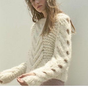 May include: A white cable knit sweater with a loose fit and openwork detailing on the sleeves.
