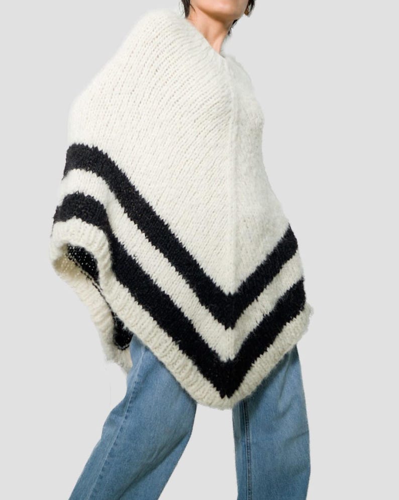 Pu&ograve; includere: Un poncho in maglia grossa color bianco sporco con due audaci strisce orizzontali nere vicino all'orlo che formano una V. Il capo &egrave; realizzato in filato spesso e testurizzato con un motivo a coste visibile, offrendo una silhouette ampia.